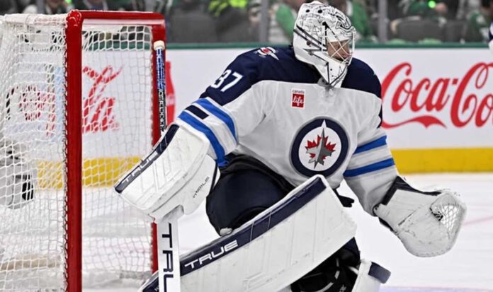 connor-hellebuyck-winnipeg-jets-trade-rumors-2026 Connor Hellebuyck in Winnipeg Jets goalie equipment defending the net with a Coca-Cola advertisement on the boards amid NHL trade rumors.