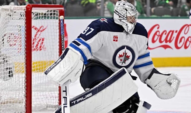 Connor Hellebuyck in Winnipeg Jets goalie equipment defending the net with a Coca-Cola advertisement on the boards amid NHL trade rumors.