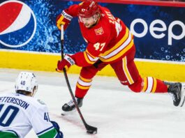 Calgary Flames forward Connor Zary (#47) skating with the puck against Vancouver Canucks Elias Pettersson in an away jersey, with a Pepsi advertisement on the boards, amid NHL trade rumors linking Zary to Vancouver.