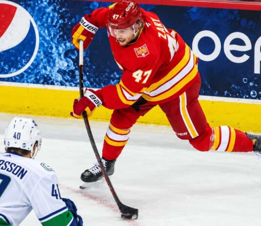 Calgary Flames forward Connor Zary (#47) skating with the puck against Vancouver Canucks Elias Pettersson in an away jersey, with a Pepsi advertisement on the boards, amid NHL trade rumors linking Zary to Vancouver.