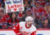Detroit Red Wings captain Dylan Larkin skates in his white away jersey as Montreal Canadiens fans cheer behind him, including a fan holding a "We Want Larkin!" sign amid NHL trade rumors.