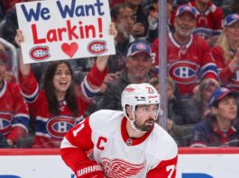 Detroit Red Wings captain Dylan Larkin skates in his white away jersey as Montreal Canadiens fans cheer behind him, including a fan holding a "We Want Larkin!" sign amid NHL trade rumors.