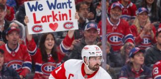 Detroit Red Wings captain Dylan Larkin skates in his white away jersey as Montreal Canadiens fans cheer behind him, including a fan holding a "We Want Larkin!" sign amid NHL trade rumors.