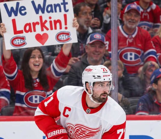 Detroit Red Wings captain Dylan Larkin skates in his white away jersey as Montreal Canadiens fans cheer behind him, including a fan holding a "We Want Larkin!" sign amid NHL trade rumors.