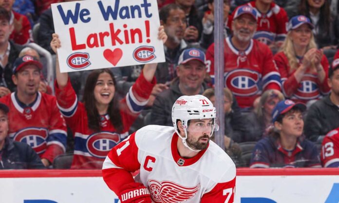 dylan-larkin-canadiens-red-wings-trade-rumors-2026 Detroit Red Wings captain Dylan Larkin skates in his white away jersey as Montreal Canadiens fans cheer behind him, including a fan holding a "We Want Larkin!" sign amid NHL trade rumors.