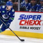 Elias Pettersson Trade Rumors: Did His Hot Start Just Change Vancouver’s Plans? Elias Pettersson skating along the boards in a Vancouver Canucks jersey beside a Super Chexx Bubble Hockey advertisement amid NHL trade rumors following his hot start to the 2026-27 season.