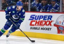 Elias Pettersson skating along the boards in a Vancouver Canucks jersey beside a Super Chexx Bubble Hockey advertisement amid NHL trade rumors following his hot start to the 2026-27 season.