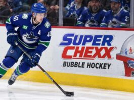 Elias Pettersson skating along the boards in a Vancouver Canucks jersey beside a Super Chexx Bubble Hockey advertisement amid NHL trade rumors following his hot start to the 2026-27 season.