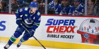 Elias Pettersson skating along the boards in a Vancouver Canucks jersey beside a Super Chexx Bubble Hockey advertisement amid NHL trade rumors following his hot start to the 2026-27 season.