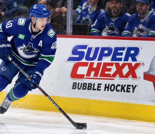 Elias Pettersson skating along the boards in a Vancouver Canucks jersey beside a Super Chexx Bubble Hockey advertisement amid NHL trade rumors following his hot start to the 2026-27 season.