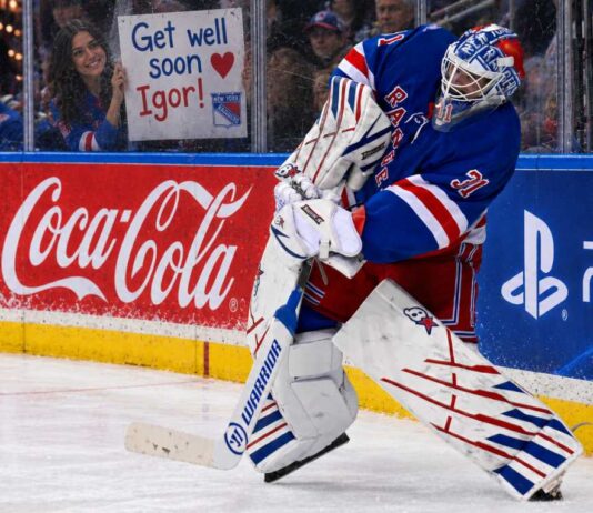 New York Rangers goalie Igor Shesterkin in action as a female Rangers fan holds a "Get well soon Igor!" sign behind the glass, following his season-ending injury. Coca-Cola and PS5 advertisements line the boards.
