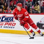 Simon Edvinsson Trade Rumors: Has Detroit Lost Another Core Piece? Simon Edvinsson skates for the Detroit Red Wings beside a Super Chexx Pro Bubble Hockey ad amid NHL trade rumors.