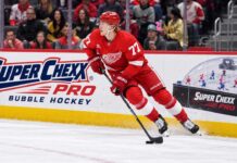 Simon Edvinsson skates for the Detroit Red Wings beside a Super Chexx Pro Bubble Hockey ad amid NHL trade rumors.