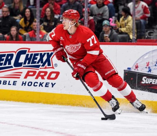 Simon Edvinsson skates for the Detroit Red Wings beside a Super Chexx Pro Bubble Hockey ad amid NHL trade rumors.