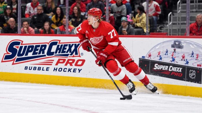 simon-edvinsson-detroit-red-wings-trade-rumors-2026 Simon Edvinsson skates for the Detroit Red Wings beside a Super Chexx Pro Bubble Hockey ad amid NHL trade rumors.